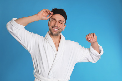 Happy young man in bathrobe and eye sleeping mask on light blue background