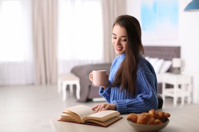 Photo of Young woman drinking coffee and reading book at table indoors, space for text. Winter season