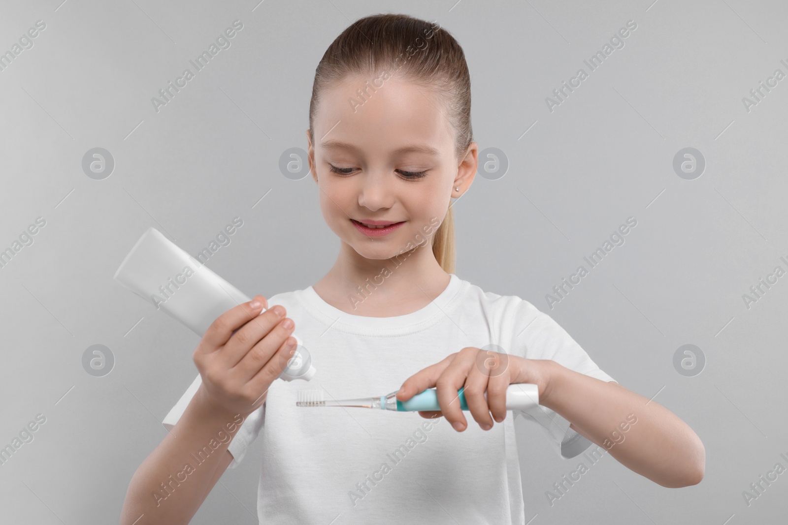 Photo of Happy girl squeezing toothpaste from tube onto electric toothbrush on light grey background