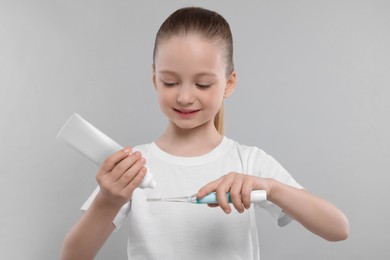 Happy girl squeezing toothpaste from tube onto electric toothbrush on light grey background