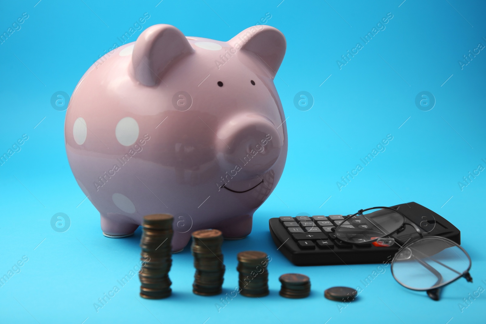 Photo of Piggy bank, stacks of coins, glasses and calculator on light blue background