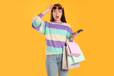 Surprised young woman with shopping bags and smartphone on yellow background. Big sale
