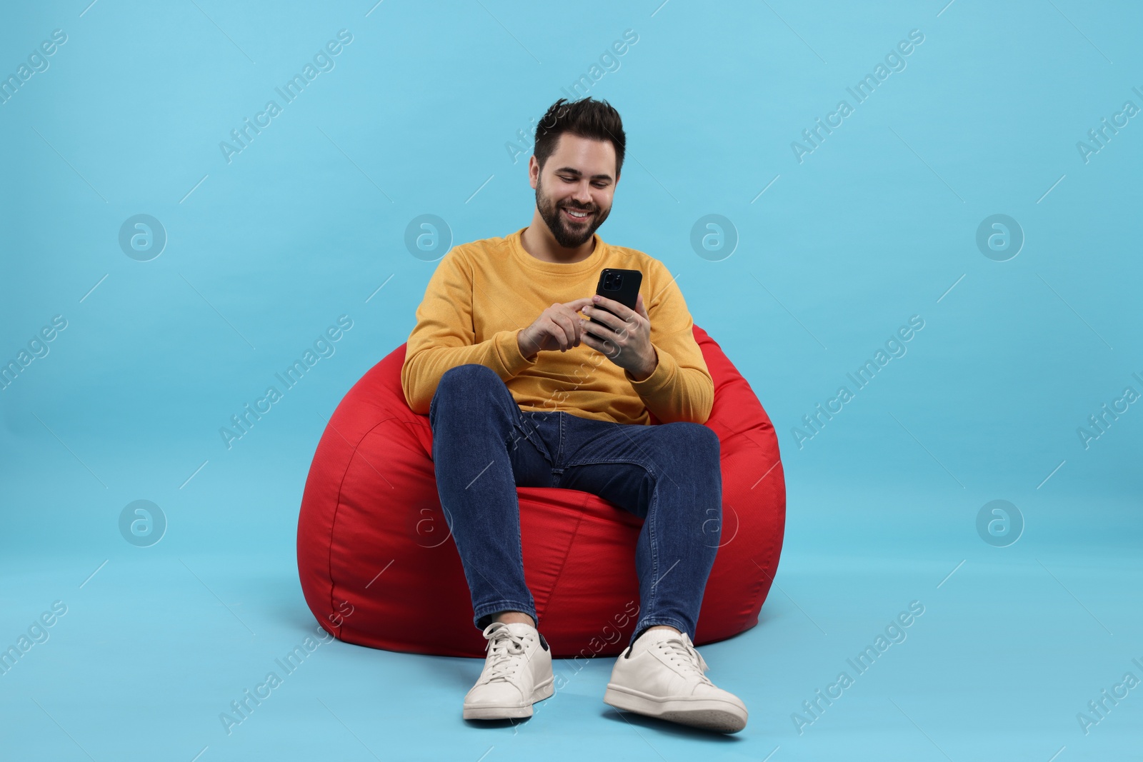 Photo of Happy young man using smartphone on bean bag chair against light blue background