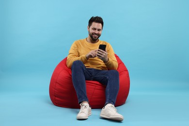 Happy young man using smartphone on bean bag chair against light blue background