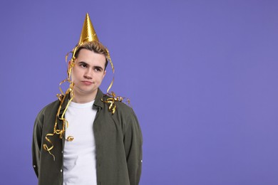 Sad young man with party hat on purple background, space for text