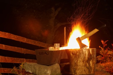 Burning firewood in metal brazier near tree stump with axe outdoors at night