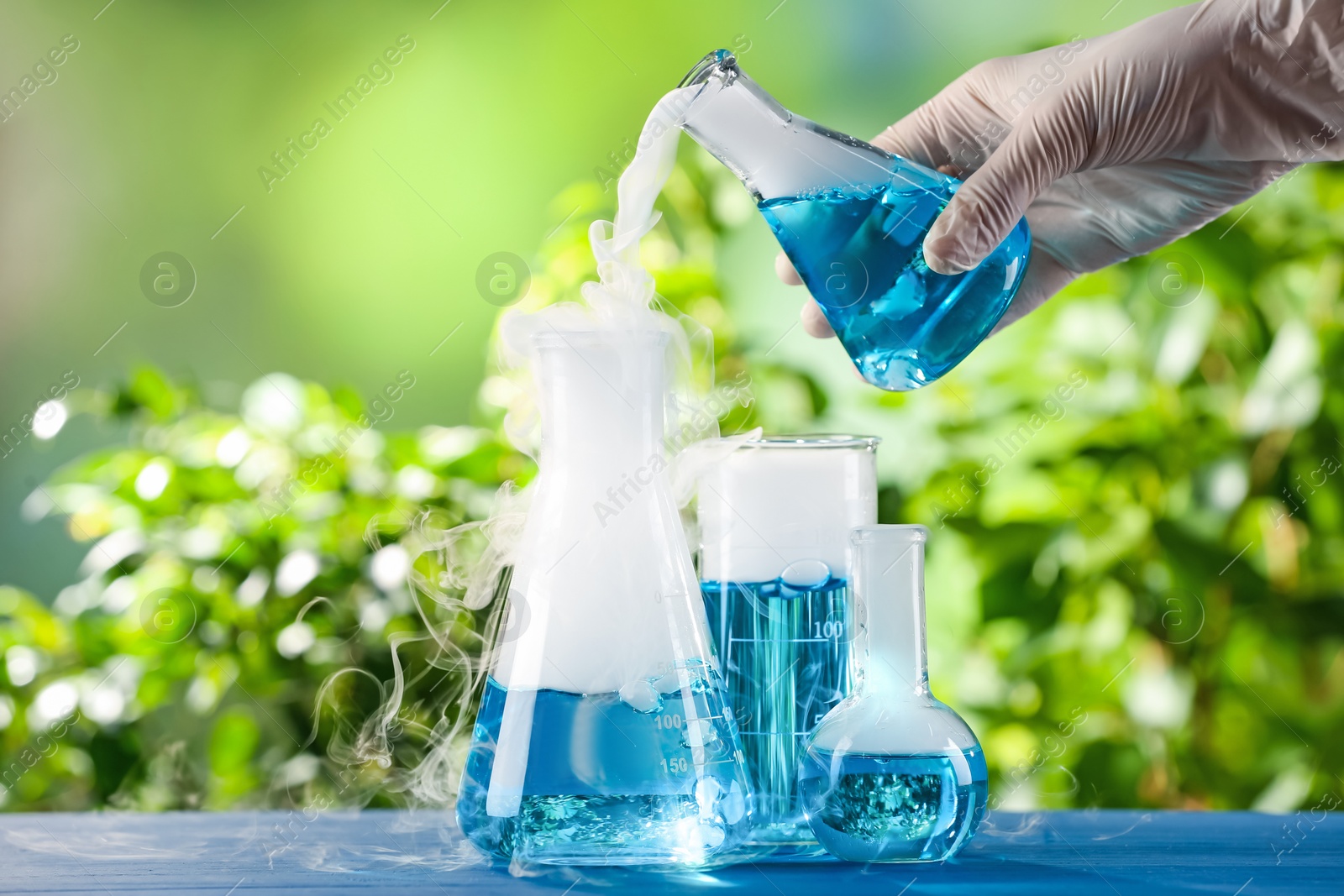 Photo of Scientist working with laboratory glassware at blue wooden table outdoors, closeup. Chemical reaction