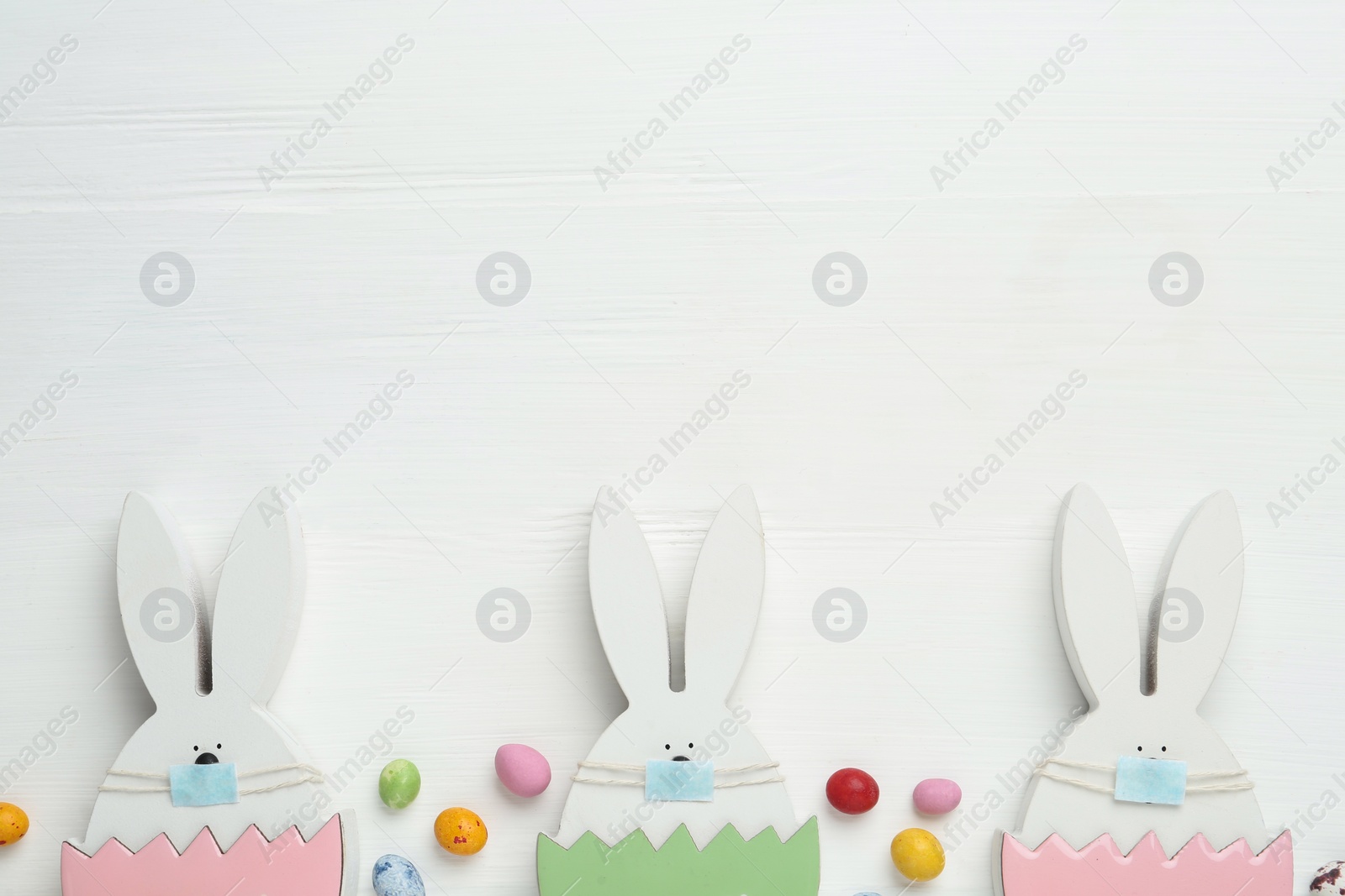 Photo of Candies and bunny figures in protective masks on white wooden table, flat lay with space for text. Easter holiday during COVID-19 quarantine