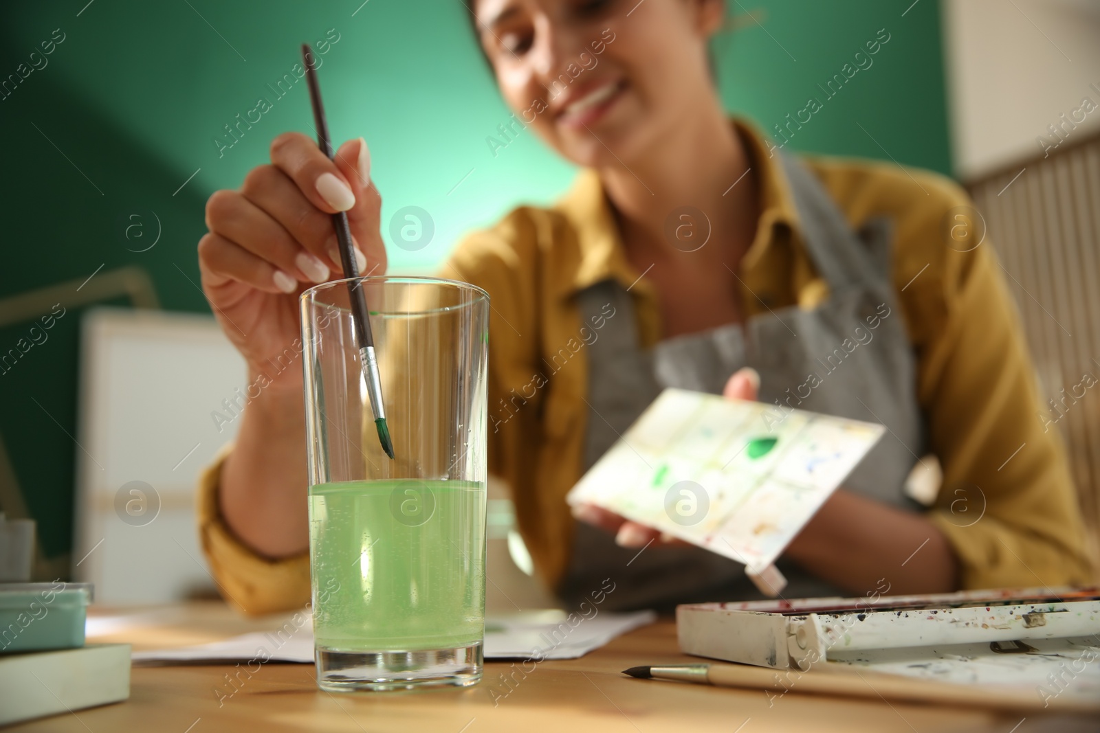 Photo of Young woman drawing at table indoors, focus on hand with brush