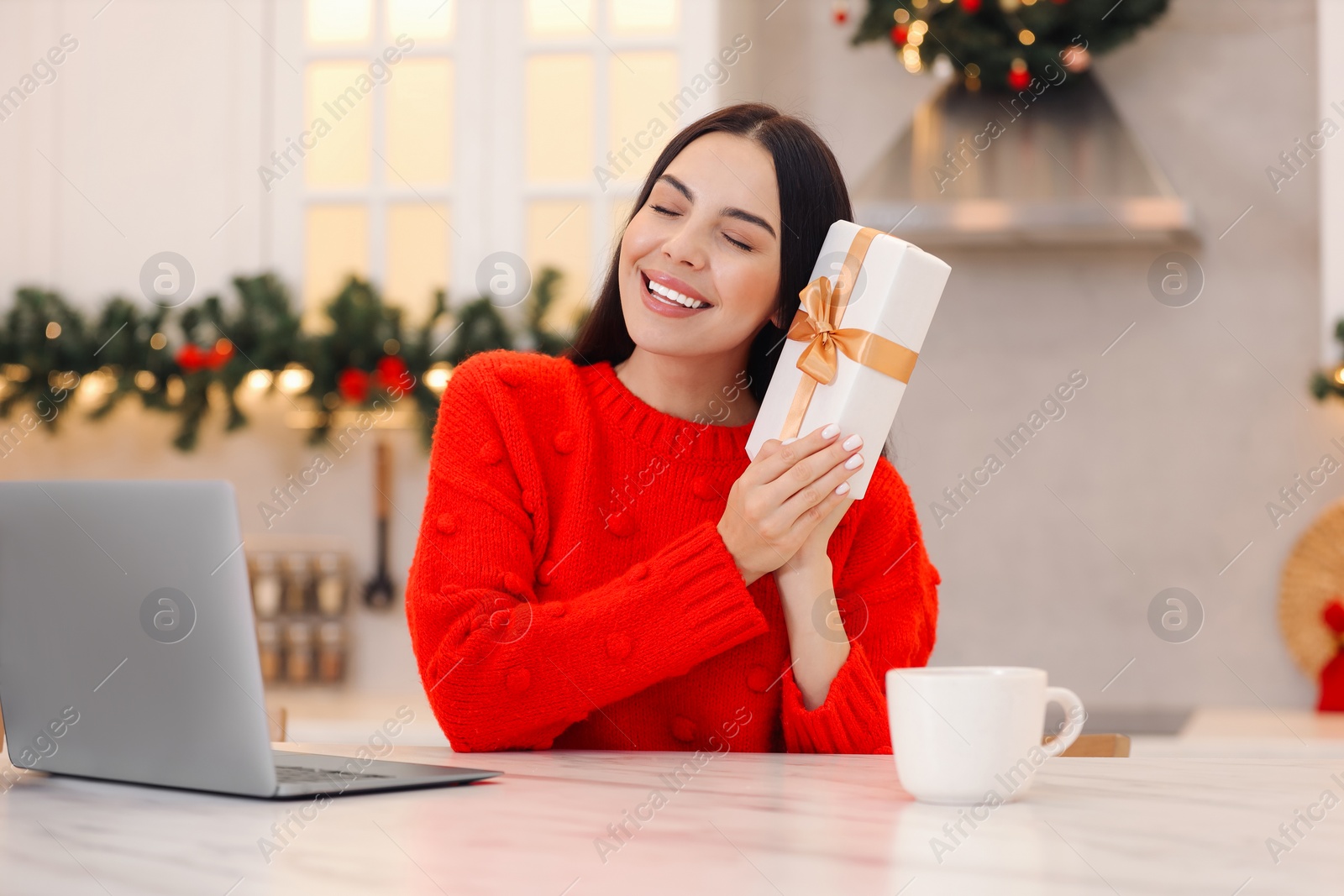 Photo of Celebrating Christmas online with exchanged by mail presents. Smiling woman with gift box during video call on laptop at home