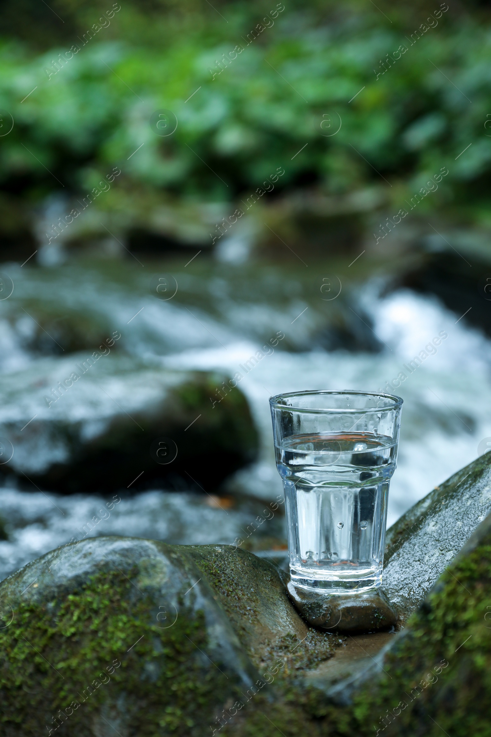 Photo of Glass of fresh water on stone near stream. Space for text