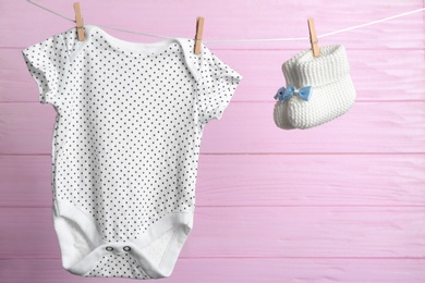 Photo of Pair of knitted booties and bodysuit on laundry line against pink wooden background. Baby accessories