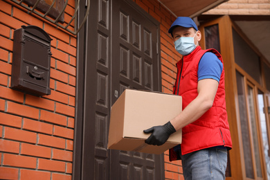 Courier in protective mask and gloves with box near house entrance. Delivery service during coronavirus quarantine