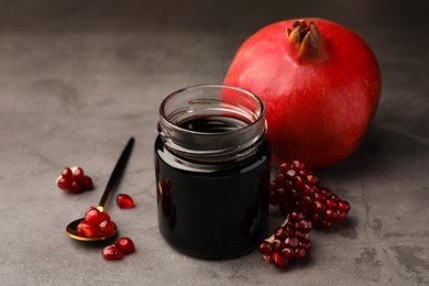 Glass jar of tasty pomegranate sauce, fresh ripe fruit and spoon on light grey table