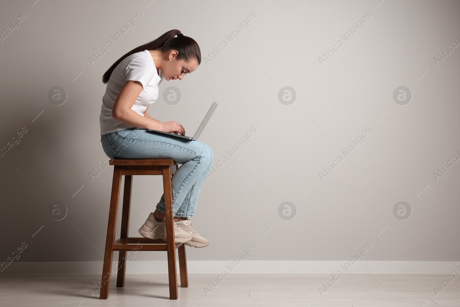 Photo of Young woman with poor posture using laptop while sitting on stool near grey wall, space for text