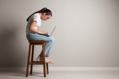 Young woman with poor posture using laptop while sitting on stool near grey wall, space for text
