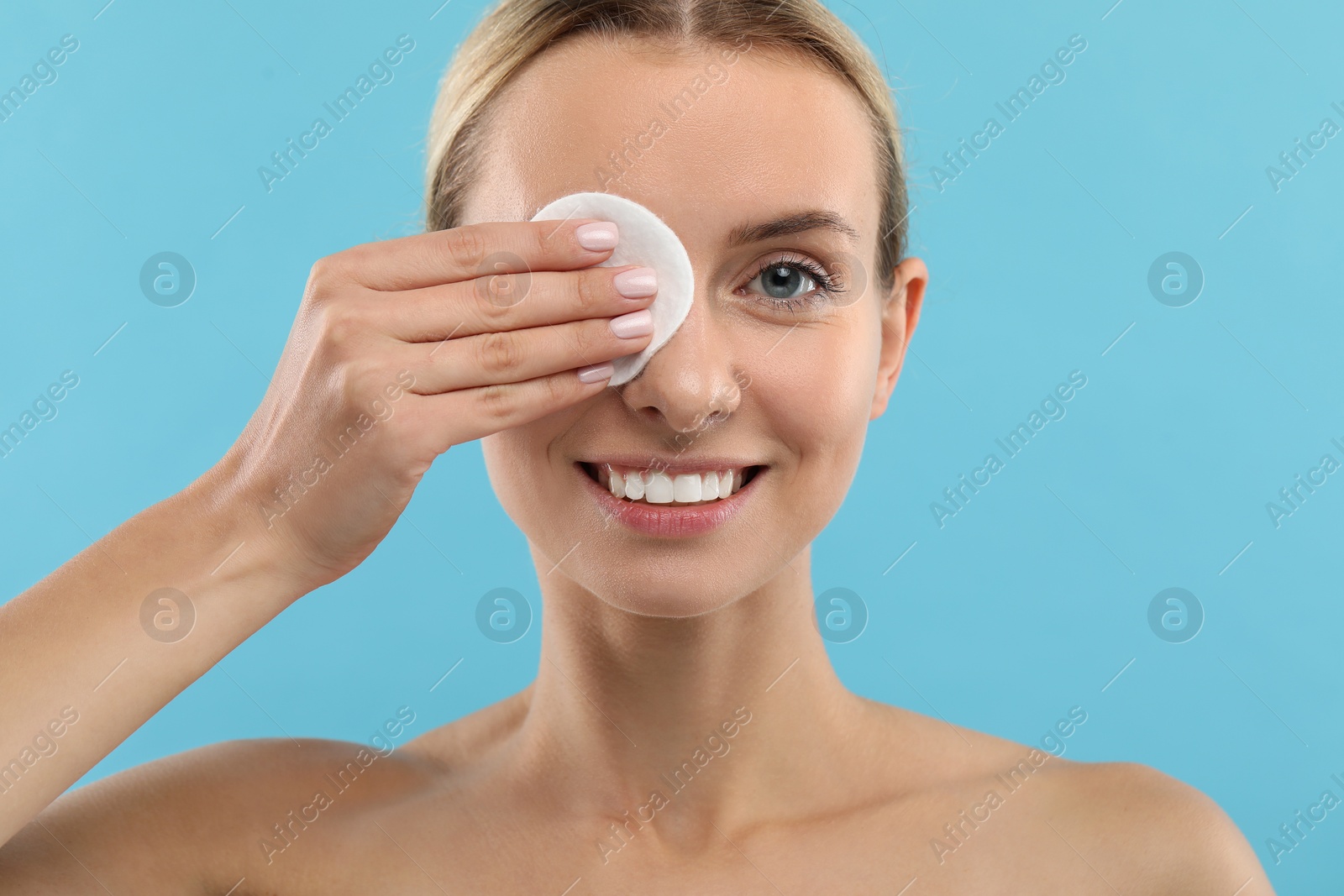 Photo of Smiling woman removing makeup with cotton pad on light blue background, closeup