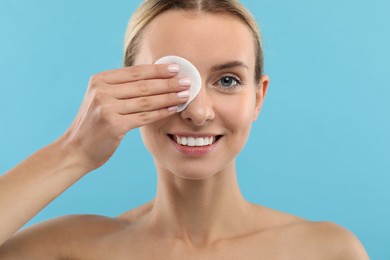 Smiling woman removing makeup with cotton pad on light blue background, closeup