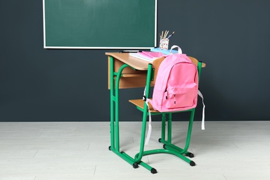 Wooden school desk with stationery and backpack near blackboard on grey wall