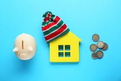 Photo of House model with warm hat, piggy bank and money on light blue background, flat lay. Paying bills concept