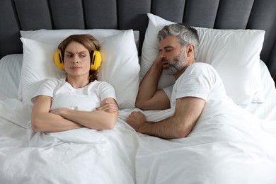Photo of Irritated woman with headphones lying near her snoring husband in bed at home