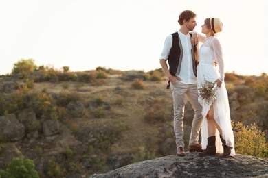 Happy newlyweds with beautiful field bouquet standing on rock outdoors