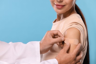 Doctor sticking plaster on woman's arm after vaccination against light blue background, closeup