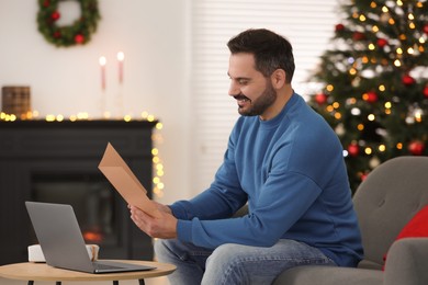 Photo of Celebrating Christmas online with exchanged by mail presents. Happy man with greeting card during video call on laptop at home