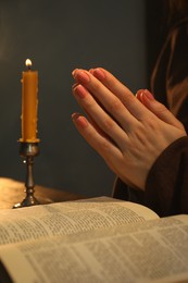 Photo of Woman praying at table with burning candle and Bible, closeup