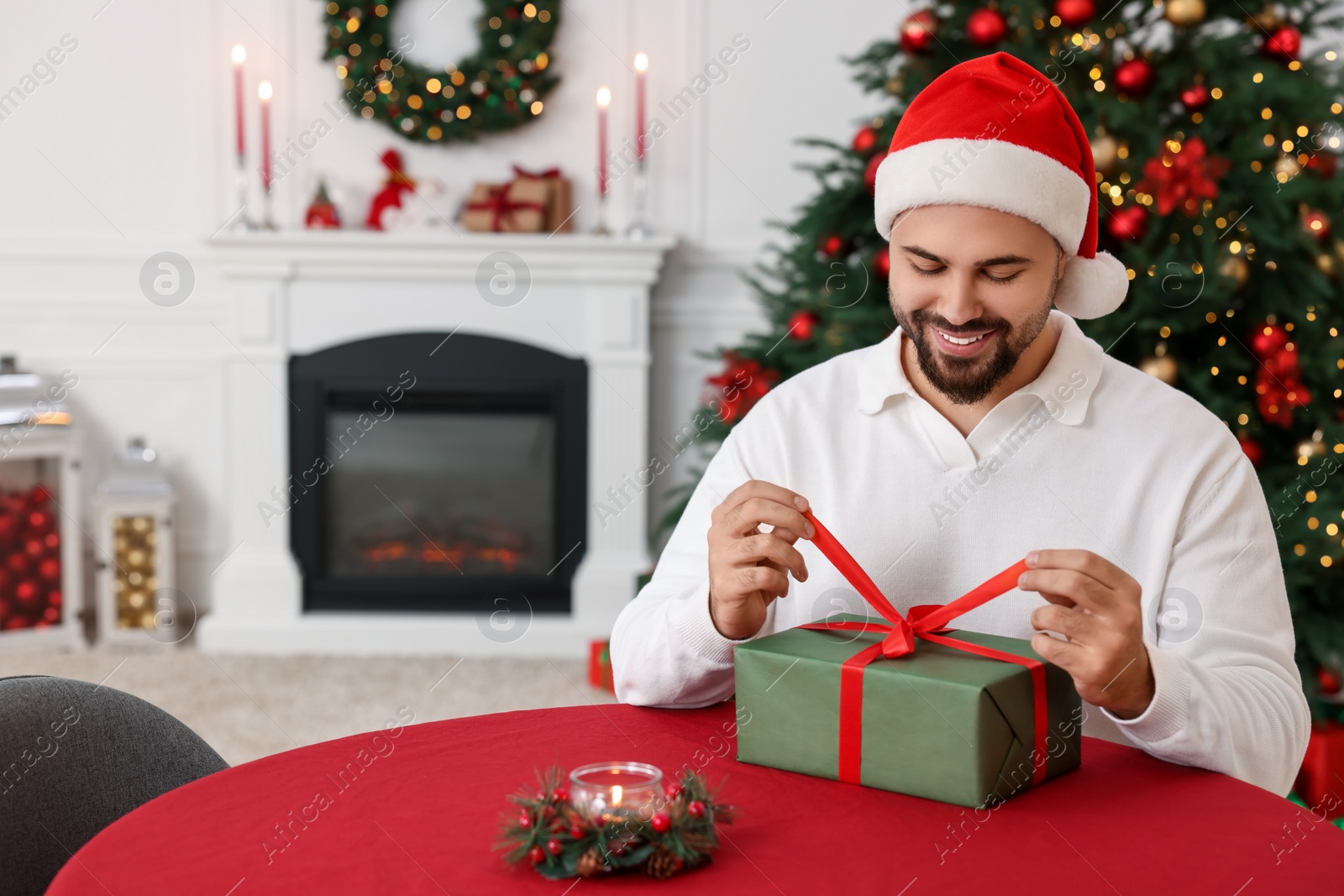Photo of Happy young man in Santa hat opening Christmas gift at table in decorated room
