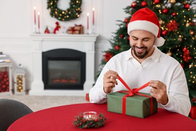 Happy young man in Santa hat opening Christmas gift at table in decorated room