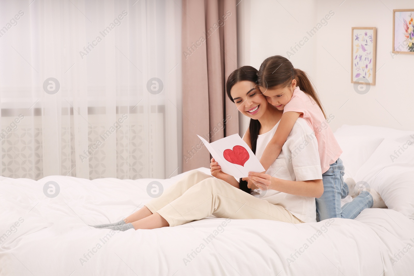 Photo of Happy woman with her daughter and handmade greeting card on bed at home. Mother's day celebration