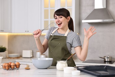 Happy young housewife with whisk having fun while cooking at white marble table in kitchen