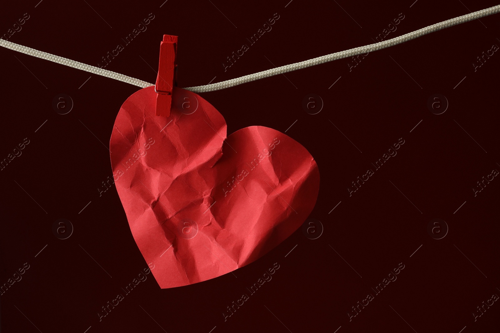 Photo of Crumpled red paper heart on rope against burgundy background, space for text. Broken heart