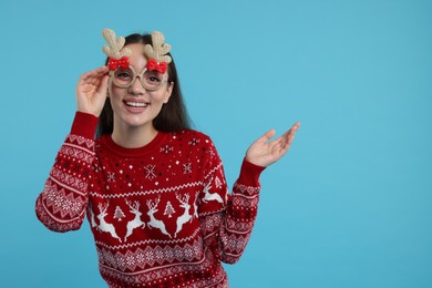 Photo of Happy young woman in Christmas sweater and funny glasses showing something on light blue background. Space for text