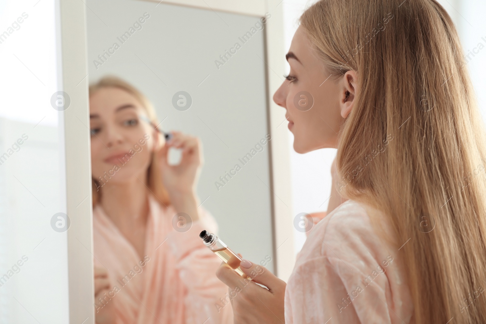 Photo of Young woman applying oil onto her eyelashes near mirror indoors