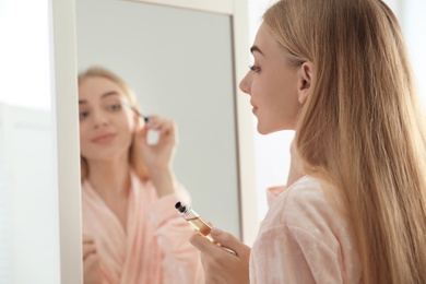 Young woman applying oil onto her eyelashes near mirror indoors