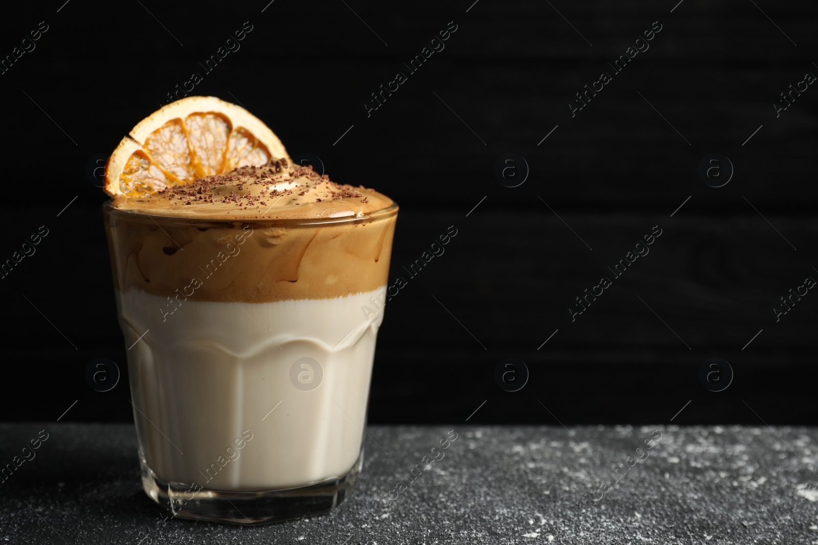 Photo of Glass of delicious dalgona coffee with dry orange and chocolate on grey textured table, space for text