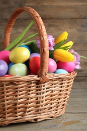 Photo of Colorful Easter eggs and tulips in wicker basket on wooden background