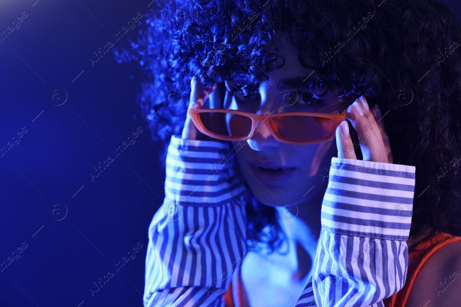 Photo of Beautiful young woman in sunglasses posing on color background in neon lights, closeup. Space for text