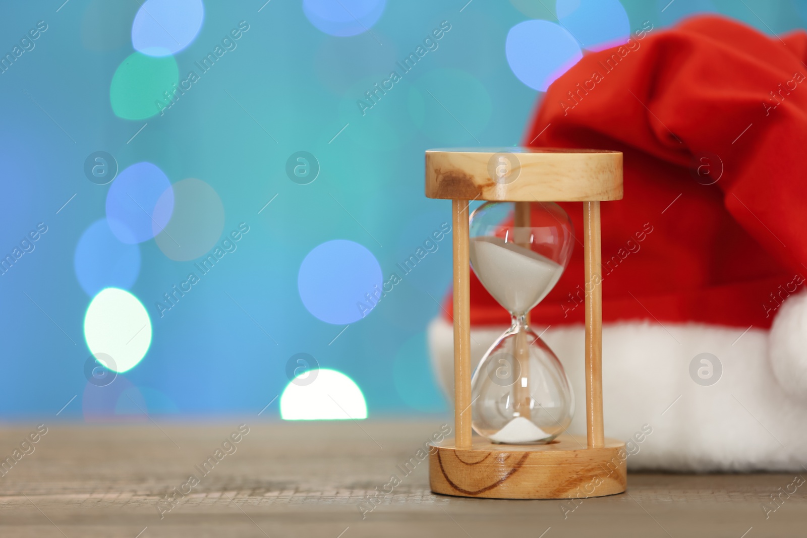 Photo of Hourglass and Santa hat on wooden table against blurred lights, space for text. Christmas countdown