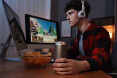 Photo of Young man with energy drink and headphones playing video game at wooden desk indoors, focus on can