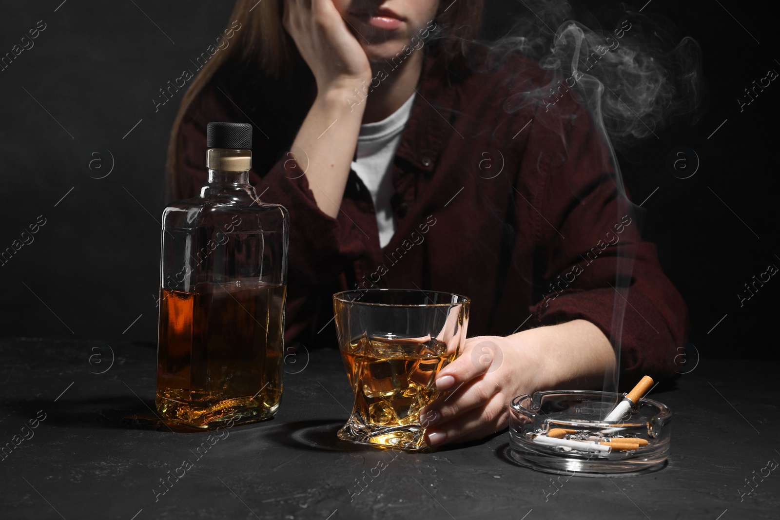 Photo of Alcohol addiction. Woman with whiskey and smoldering cigarettes at dark textured table, closeup