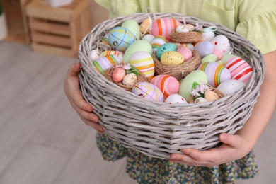 Photo of Little girl holding wicker basket full of Easter eggs indoors, closeup