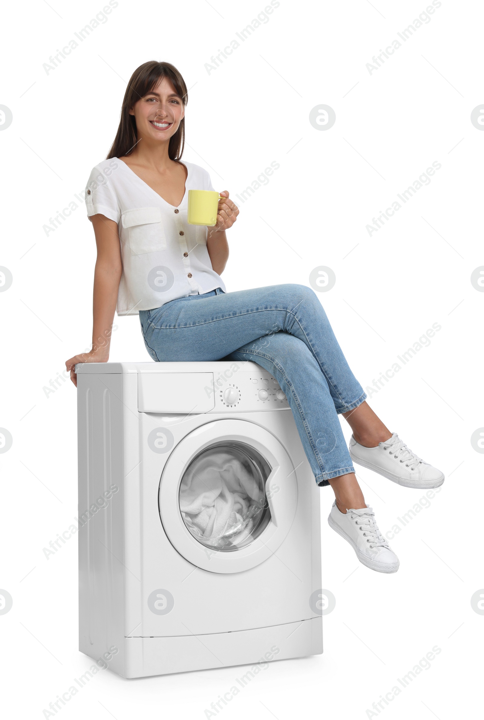 Photo of Beautiful woman with cup of drink sitting on washing machine against white background