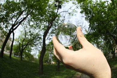 Photo of Beautiful green trees outdoors, overturned reflection. Man holding crystal ball in park, closeup. Wide-angle lens