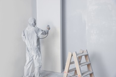 Photo of Decorator in uniform painting wall with sprayer indoors, back view