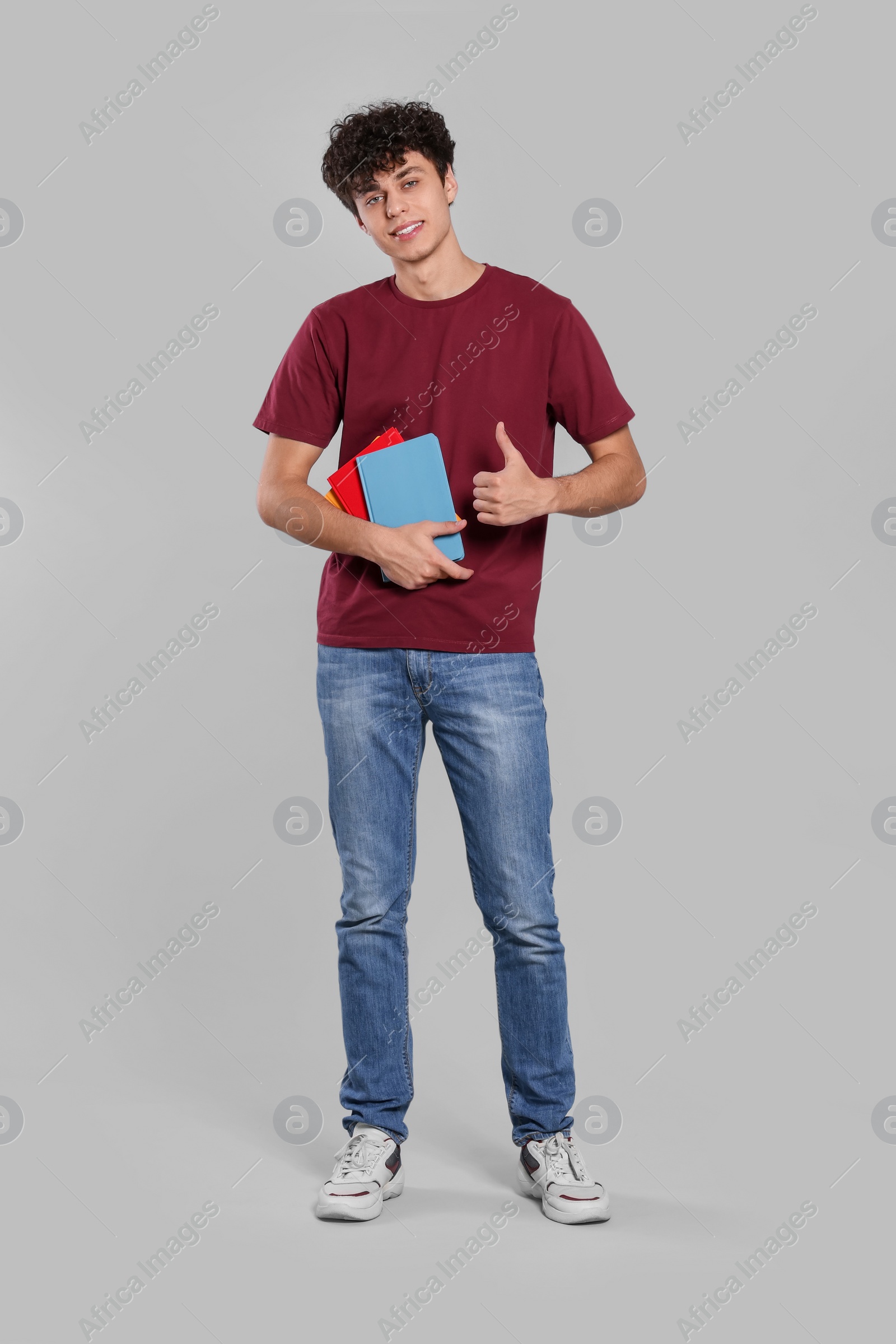 Photo of Handsome young man with books showing thumb up on light grey background