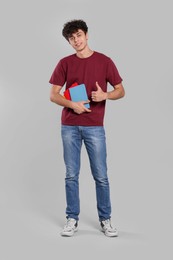 Photo of Handsome young man with books showing thumb up on light grey background