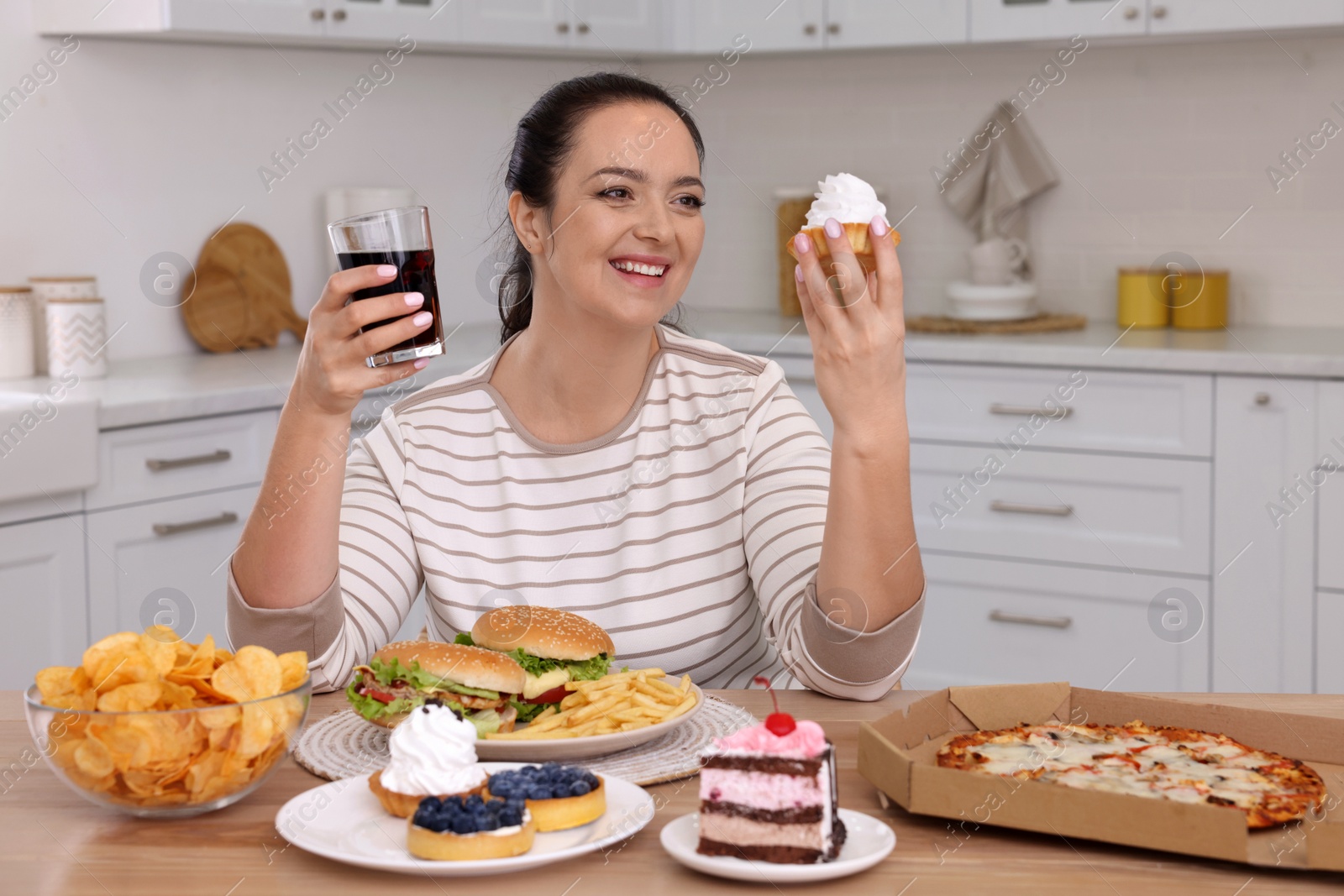 Photo of Happy overweight woman with glass of cola and cake in kitchen. Unhealthy food